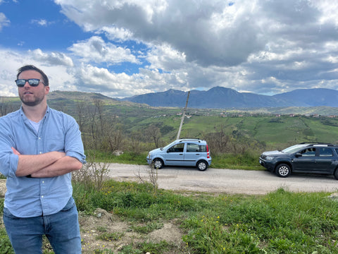 Man standing in a vineyard in Campania, Italy, surrounded by rows of green grapevines, with rolling hills and mountains visible in the background.