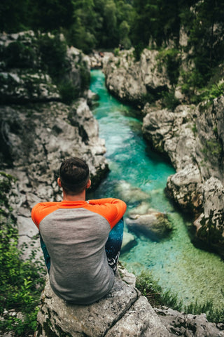 Mann in einem grau-orangen Merinoshirt schaut auf einem Felsen sitzend auf einen Bergfluss