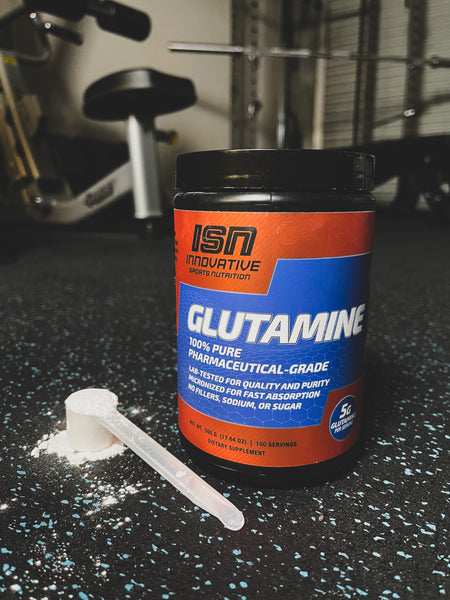 ISN orange and blue label glutamine bottle next to a scoop of white powdered glutamine on a black floor with blue specks. 