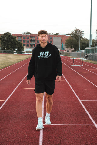 Man walking on a red running track in a black sweatshirt and black shorts. 
