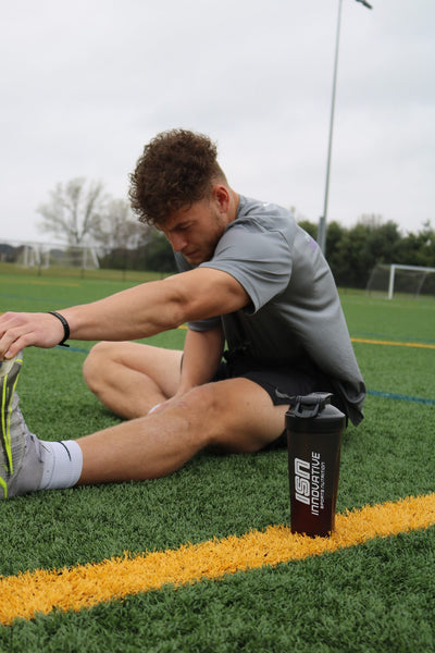 A young, male soccer athletic stretching his hamstring on a soccer field with a black ISN shaker bottle next to him. 