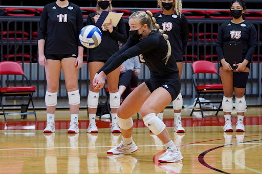 Blonde volleyball athlete dressed in all back uniform wearing a facial covering passes a volleyball during a volleyball match. 