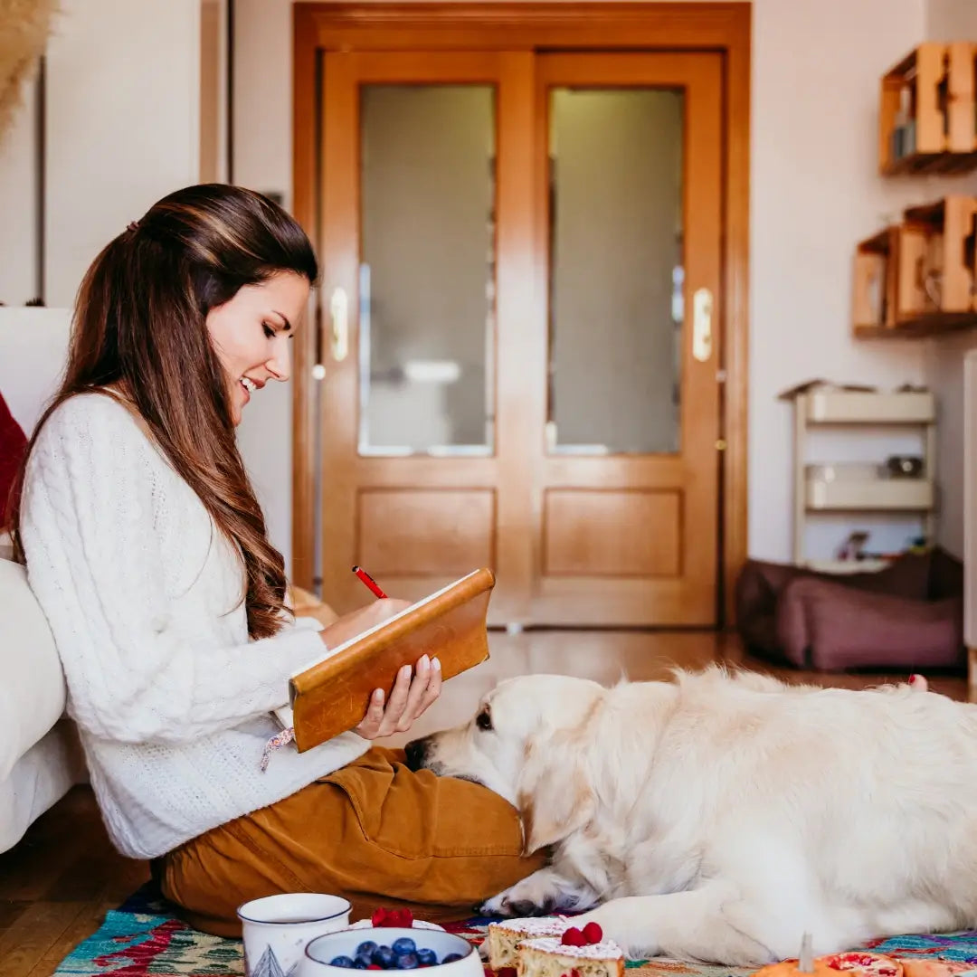 A person sits on a colorful rug, writing in a notebook, with a golden retriever resting its head on their lap and a small picnic spread nearby.