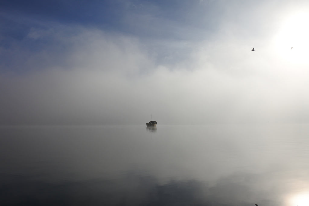 A small boat sits perfectly still on a glassy lake, with big tufts of fog on the horizon and the sun peeking through, some birds flying silently in the morning sun.