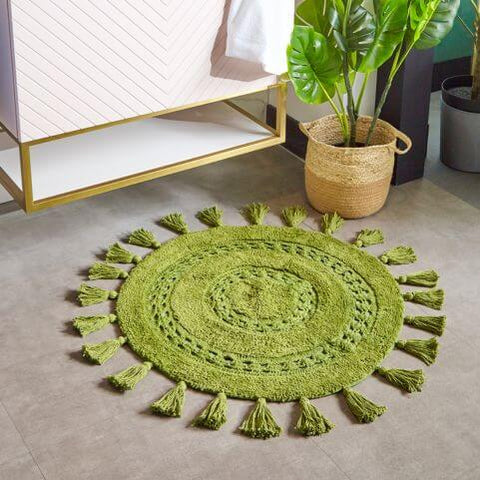 A circular green bath mat with intricate mandala weaving and textured tassels around the border.