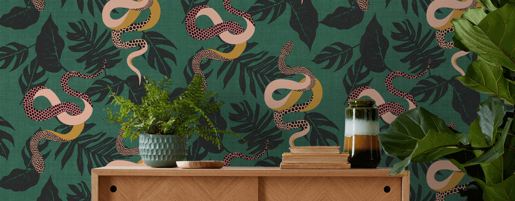 A snake patterned green wallpaper behind a light wood sideboard with a plant, books and other decorative items
