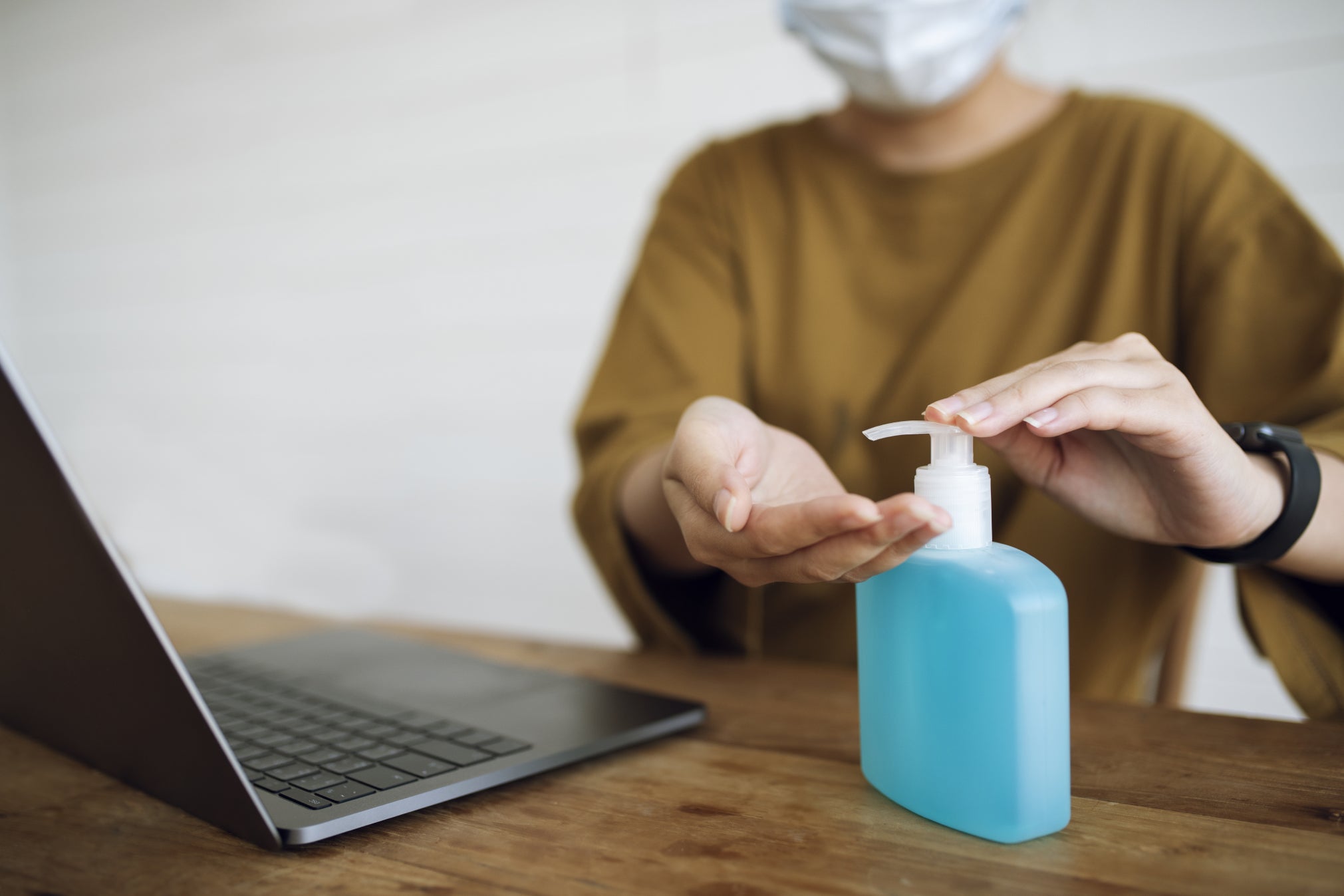 Women with face mask on sanitizing her hands while sat at desk on laptop