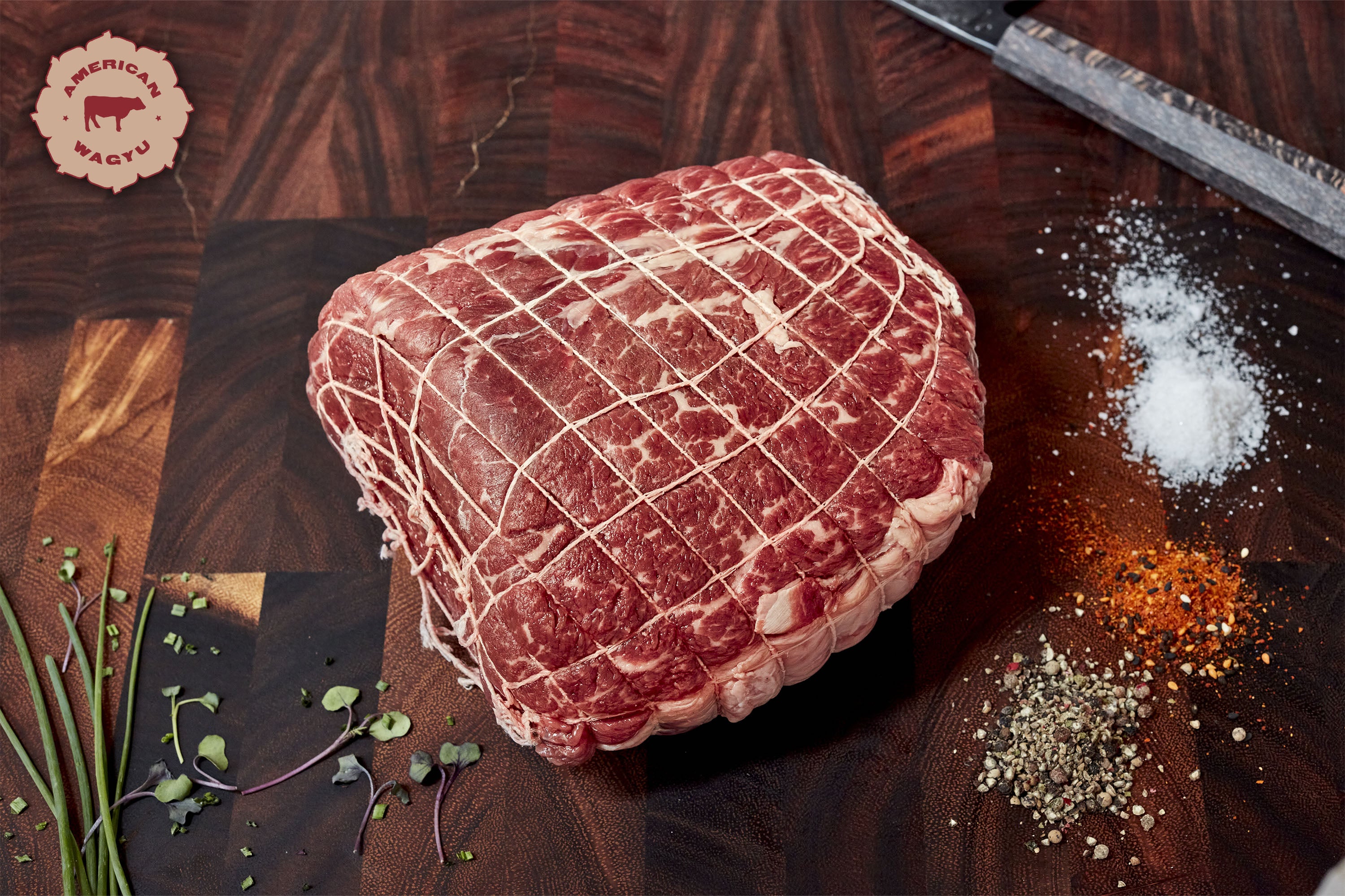 Raw, netted Wagyu beef roast on a wooden cutting board with spices and herbs nearby.