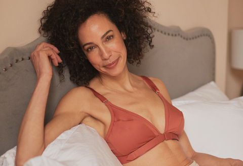 Beautiful mature woman reclining in bed, playing with her hair while softly smiling at camera, wearing the Springrose adaptive bra in terracotta