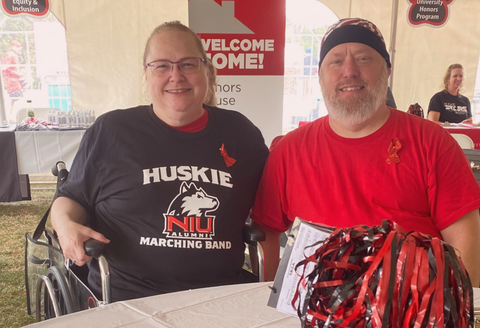 Jill and her husband smiling happily at camera. Jill is sitting in her wheelchair and wearing a Huskies shirt, while her husband is wearing a red shirt. They're at a reunion weekend.