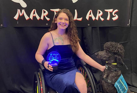 Brie smiling widely with her award, while in her wheelchair, with her dog next to her