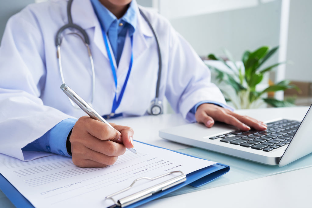 female doctor writing down assessment for patient with laptop in front of them