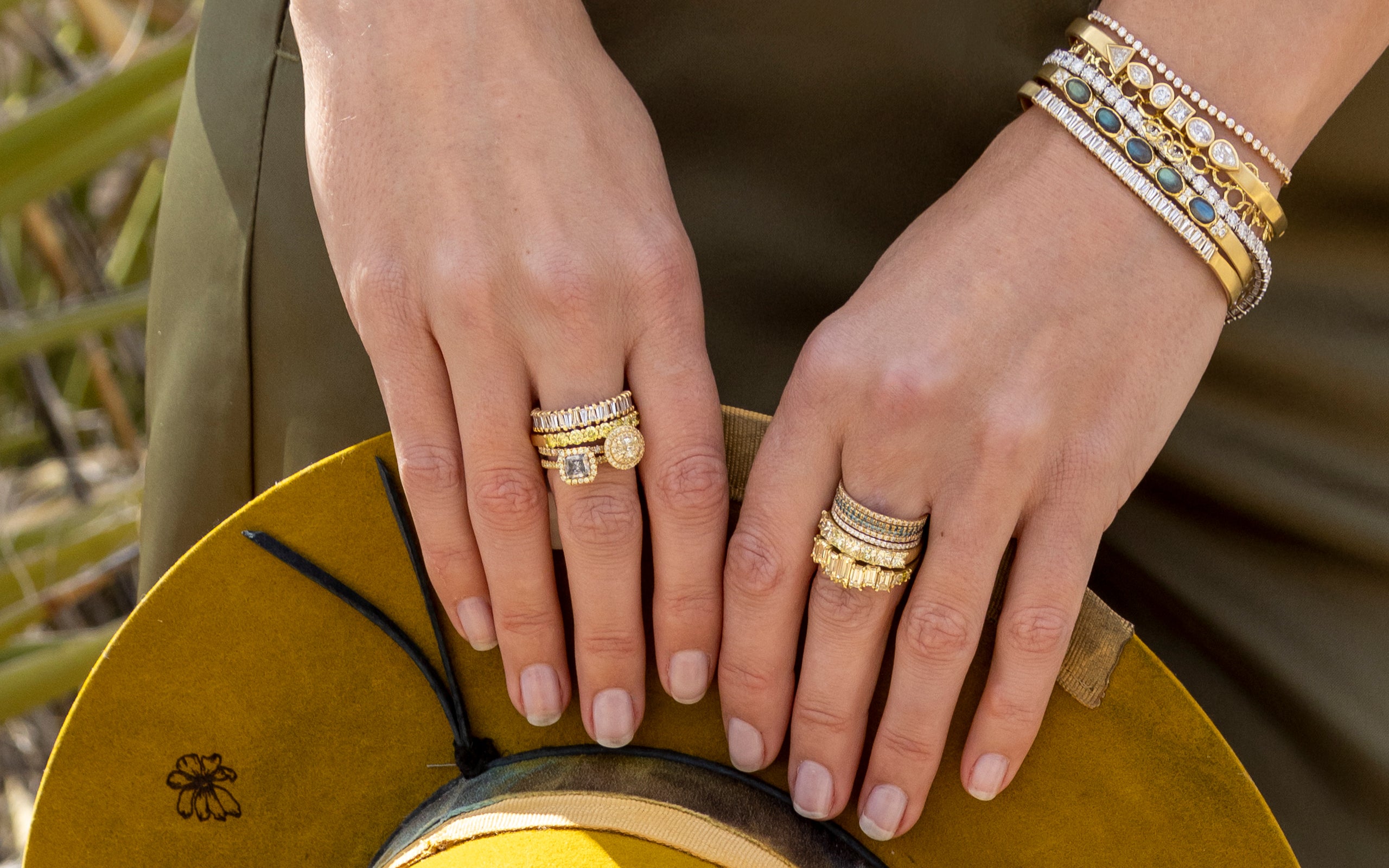 Closeup of female hands wearing diamond, yellow diamond, grey diamond, labradorite, chic designer gold jewelry; holding designer yellow hat; phone, text 323-404-2959 for shopping help.