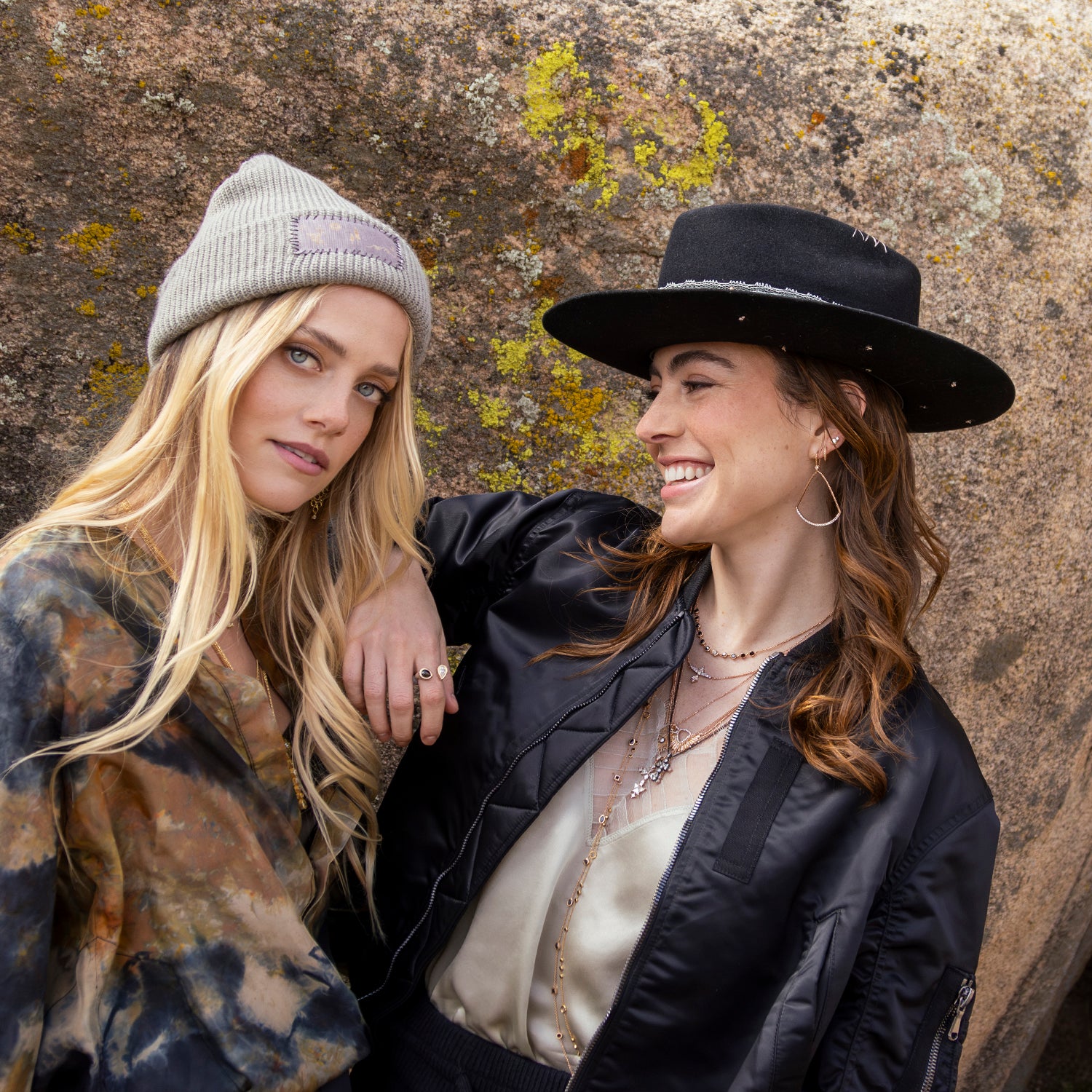 Woman in black hat, woman in designer beenie laughing in front of rock while wearing diamond jewelry, silk apparel, designer bomber jacket; phone 323-404-2959 for shopping help.