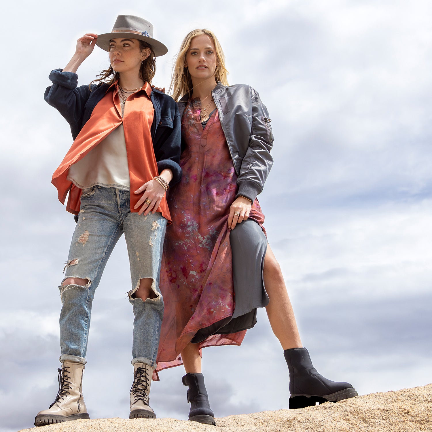 Pair of women standing atop a rock in the breeze while wearing Dominique Cohen silk apparel, 18k rose gold and black gold diamond jewelry, and a limited edition hat; shopping assistance call text 323-404-2959