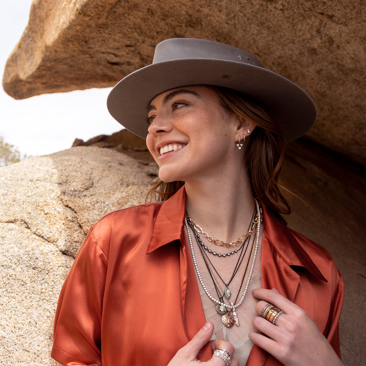 Closeup of smiling woman in Dominique Cohen black gold and rose gold diamond jewelry, silk shirt, designer hat while standing beneath rock outcropping; shopping assistance call text 323-404-2959