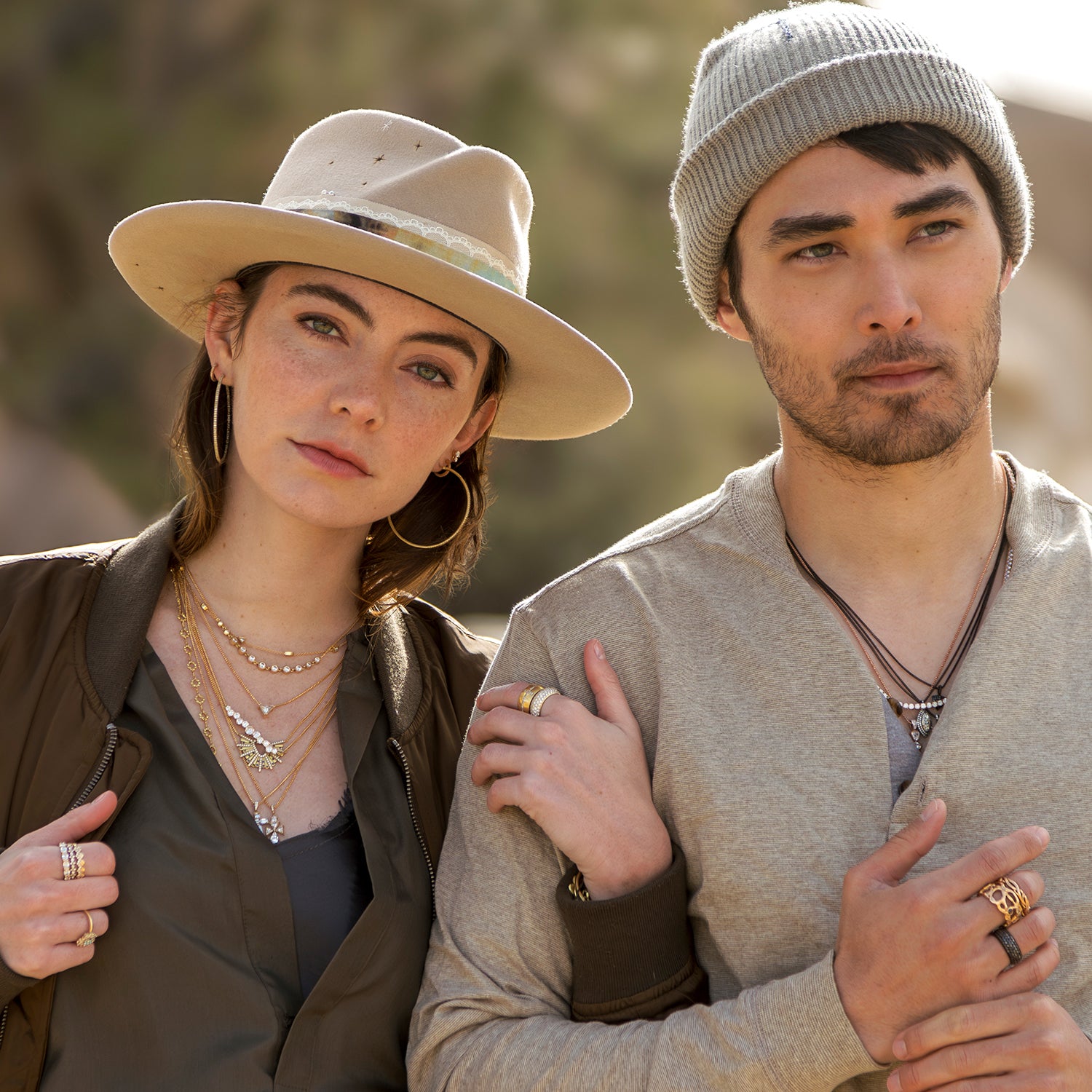 Laid back luxe arm in arm woman, man wearing black gold, yellow gold diamond jewelry, designer bomber jacket, designer hats while in Joshua Tree national park; phone 323-404-2959 for shopping help.