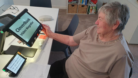 Beverley at her home desk using the Clover Book Pro with a smile. The Clover 6 is on the table next to her.