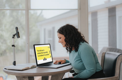 Woman at her desk leaning forward to read the laptop screen. OrCam Read 3 is set up in a stand next to the latop, reading a book.