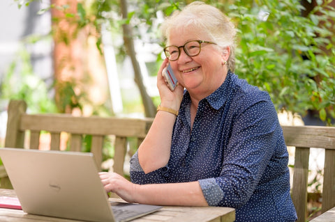 Smiling woman on phone with laptop in front of her, sitting outside