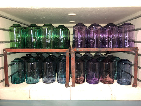 Shelves neatly lined with collections of glassware, featuring rows of green and purple tumblers turned upside down on rustic metal stands, presenting a colorful and organized display within a cabinet or pantry.