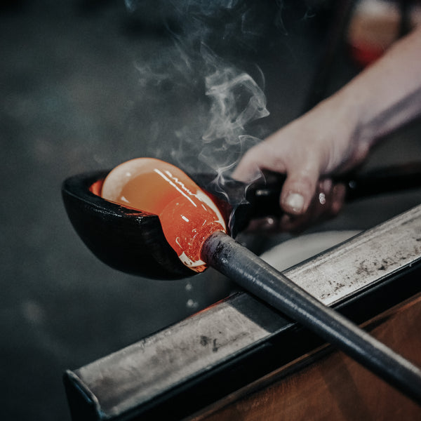 Steam rises from a molten glass form as it's cradled in a shaping tool, with the glassblower's hands carefully guiding the process. The vibrant orange hue of the hot glass stands out against the dark, blurred background of the glassblowing studio.