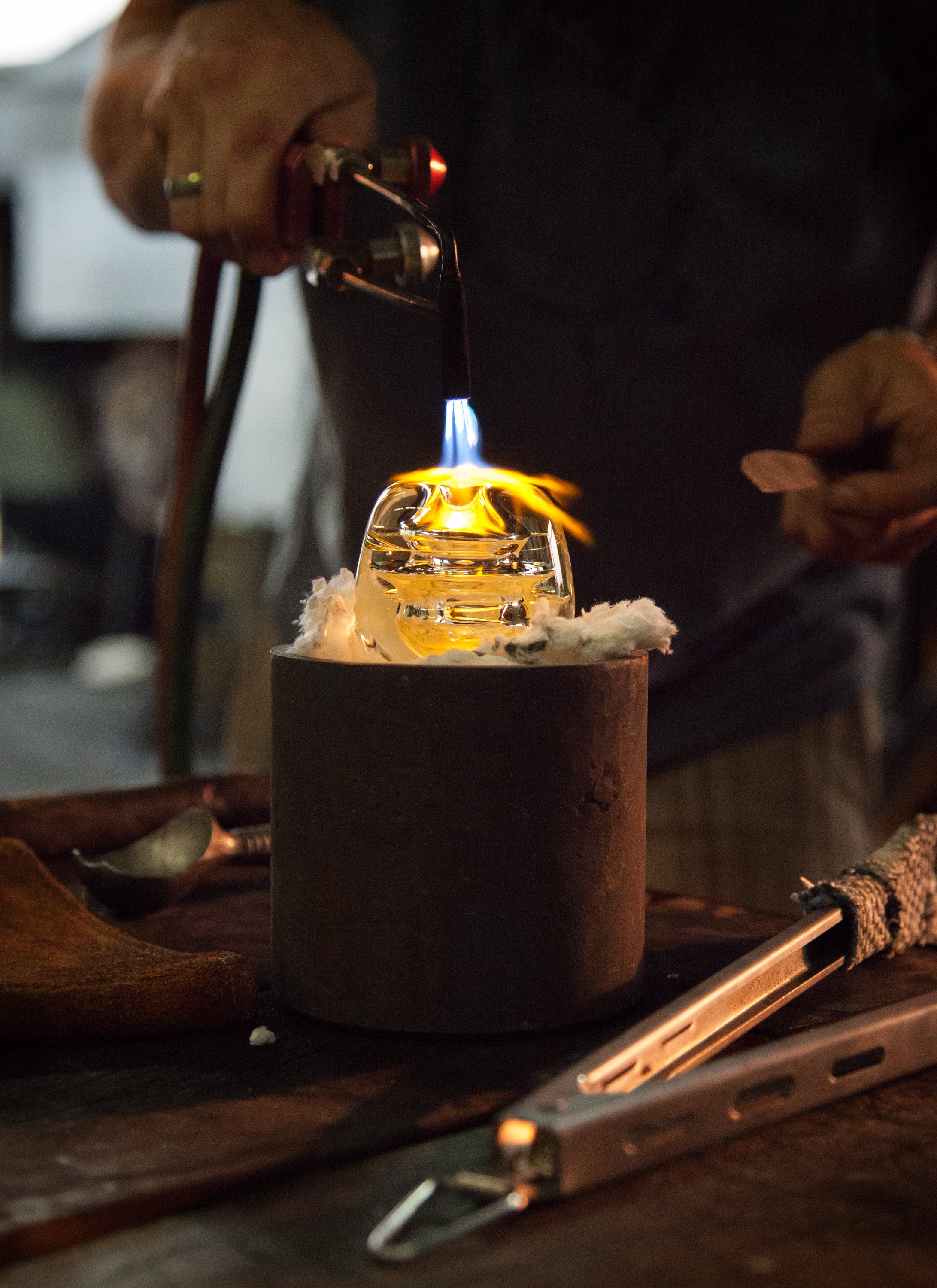 Artisan glassblowing process captured in action, showcasing the skilled hand of a craftsman using a blowtorch to shape a glowing piece of molten glass on a marver. Tools of the trade, like tongs and a steel table, frame the scene, emphasizing the blend of artistry and precision in handcrafted glasswork.