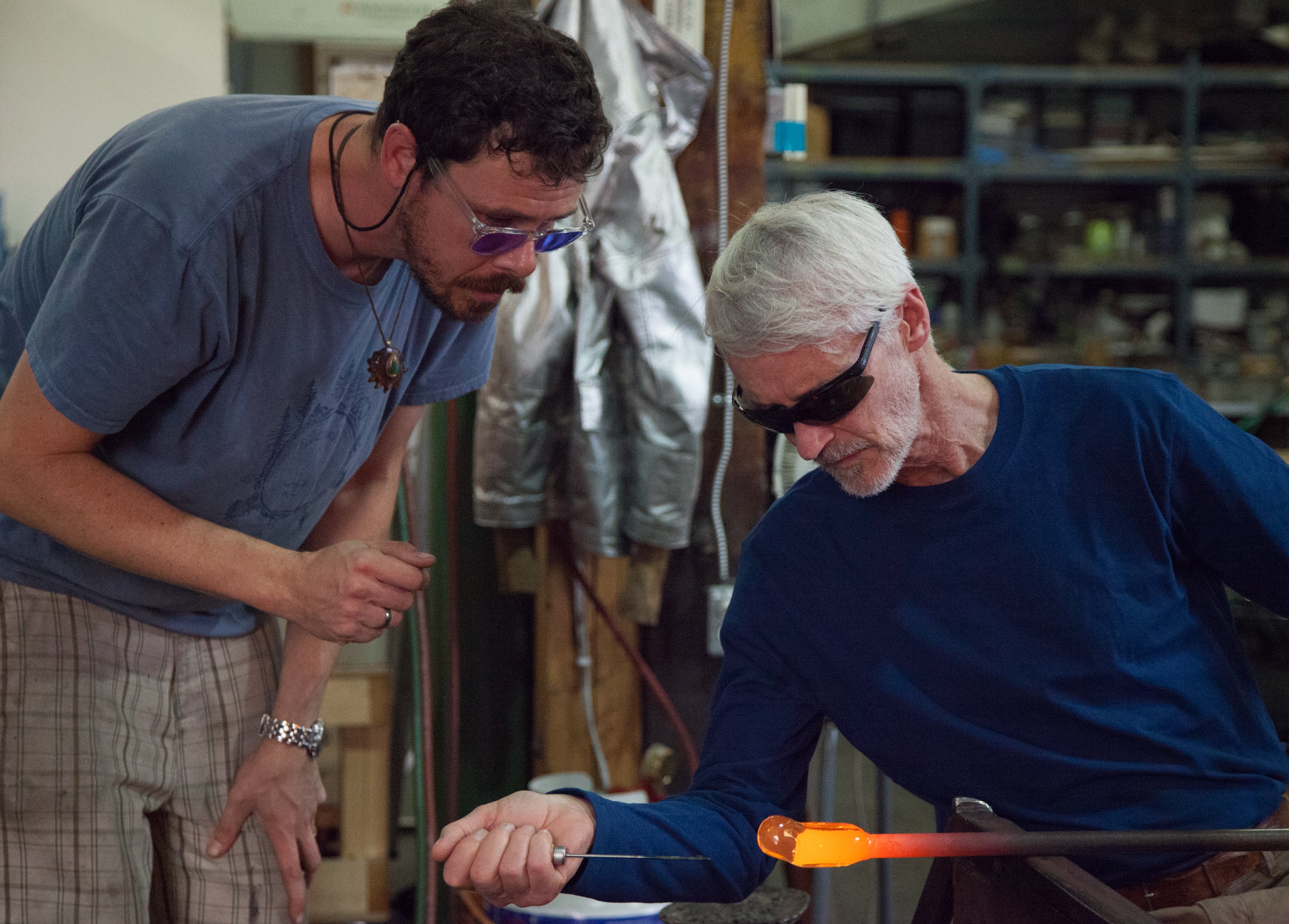 A concentrated glassblowing session in progress, featuring two individuals in safety glasses focused on shaping a hot, glowing piece of molten glass. The elder artisan holds the glassblowing pipe, shaping the viscous material, while the younger one observes closely, set against a backdrop of a cluttered workshop filled with tools of the trade.
