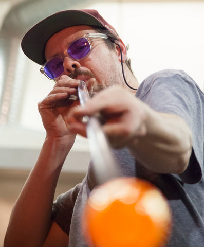 Close-up of a glassblower in concentration, wearing purple safety glasses and a cap, as they shape a glowing, molten glass form with a blowpipe, highlighting the skill and focus required in the glassblowing craft.