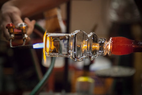 A glass artist meticulously shapes a transparent glass object with a torch, focusing the flame on a specific area. The fiery glow accentuates the craftsmanship involved in glasswork against a blurred workshop background.