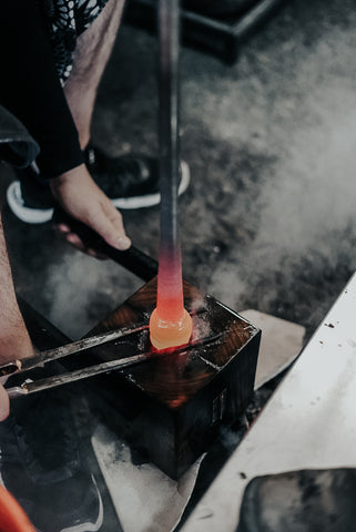 A detailed look at the art of glassblowing, where hands skillfully shape a glowing molten glass piece with tongs over a wooden mold, the intensity of the craft evident in the focused glow and contrasting workshop environment