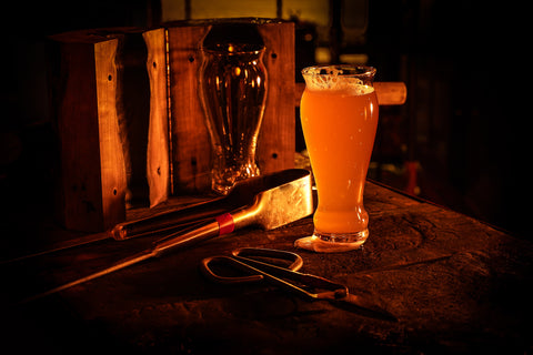 A moody and atmospheric photo capturing the essence of a glassblower's workshop, with a tall beer glass overflowing with frothy liquid on the foreground anvil. Glassblowing tools rest alongside, and a wooden mold with a vase silhouette in the dark, glowing background adds to the artisanal vibe