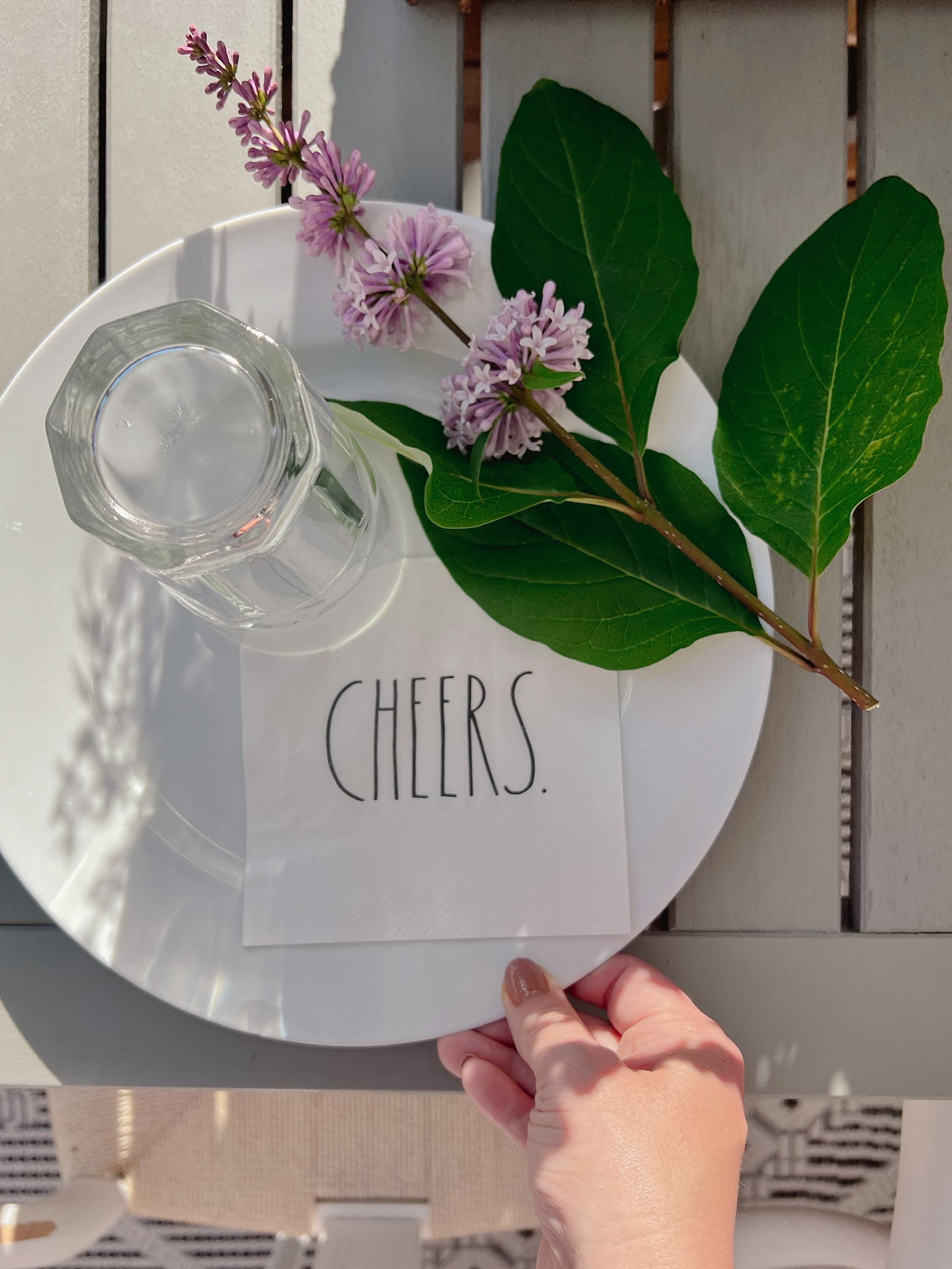 Rae Dunn napkins paired with white dishes on a  Ikea Bondholmen Dining Table with white wishbone chairs set for an outdoor dinner party . Petunia and herb planter decorates the outdoor dining table