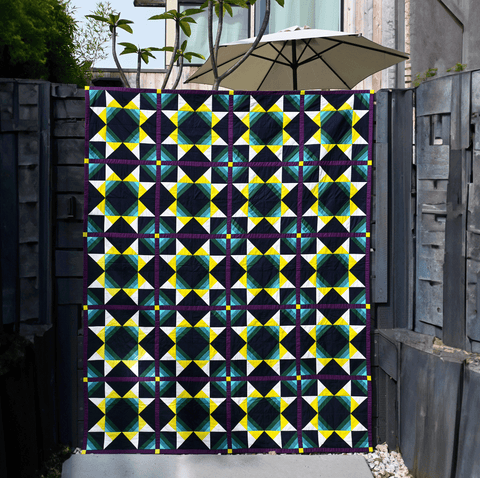 The Solar Wind quilt hangs on a brown fence. The quilt is dark navy with teal, purple, green, white, and bright yellow shapes.