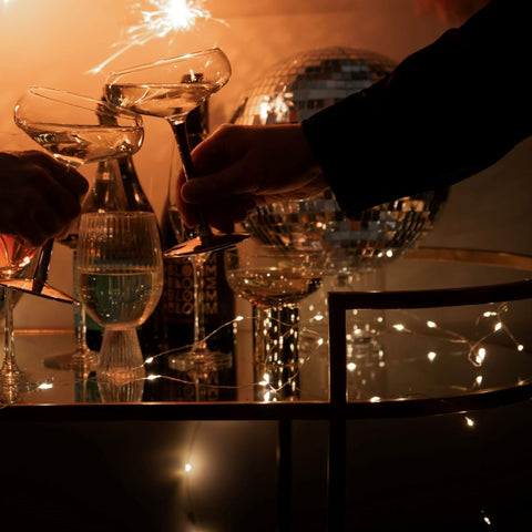 two hands clinking champagne glasses over a bar cart with fairy lights