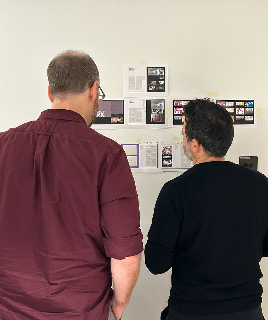 Two men stand with their backs to the camera, discussing design work in progress on the wall.