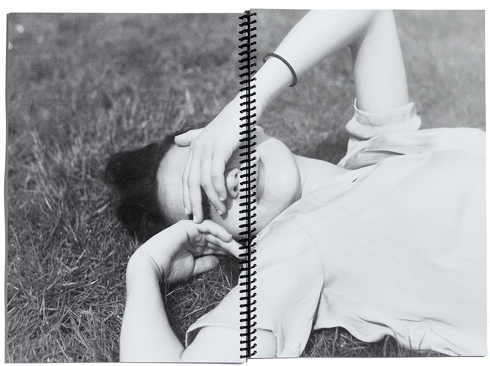 Double page spread, with spiral bning at entre, showing a black and white image of a woman lying on grass shielding her eyes from the sun.