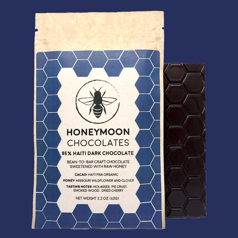 A photo of our 85% Haiti Dark Chocolate Bar next to its packaging which is a pale cream and dark indigo color