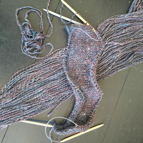 A slightly shiny handspun yarn being knit into a rectangle. The yarn appears to be dark grey but actually has lots of silver, dark teal and burgundy throughout.