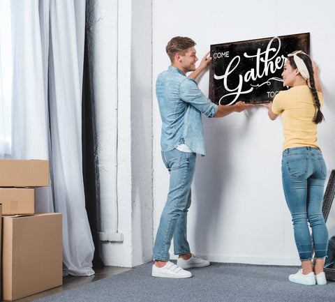 Newlyweds hanging up custom wall art in their new home. Moving boxes are in the background and couple is hanging a black sign with white lettering that says "gather".