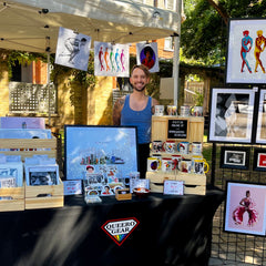 Eric Van Giessen standing behind a Queero Gear booth at the Toronto Queer Market. On the table infront of him are art prints in wooden bins, coffee mugs with art illustrations and stickers lined up on a shelf. To his right is a grid wall with framed prints. 