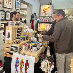 Eric Van Giessen standing behind a Queero Gear booth at The Tiz The Szn market run by The Welcome Market and Of Sorts. In front of him are art prints in wooden crates and coffee mugs on shelving. Behind him is a gallery display of framed art prints and there are two men shopping at the booth. 