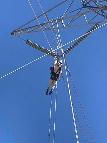 A utility lineman ascends a rope to access a hot plate on a transmission tower in rough terrain