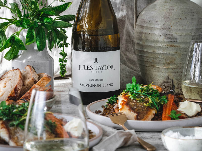 Bottle of Jules Taylor Sauvignon Blanc wine on table with food, wine, plants, urn.