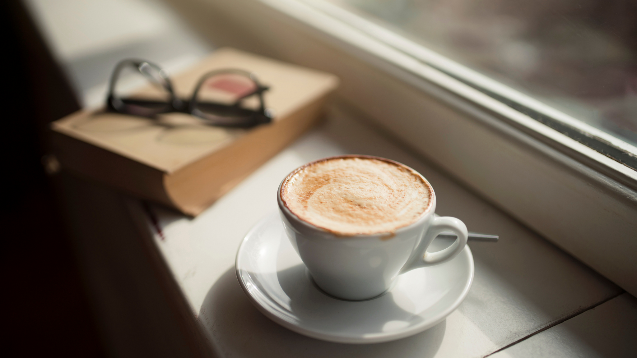 Photo of a latte on a windowsill next to a book with glasses on top