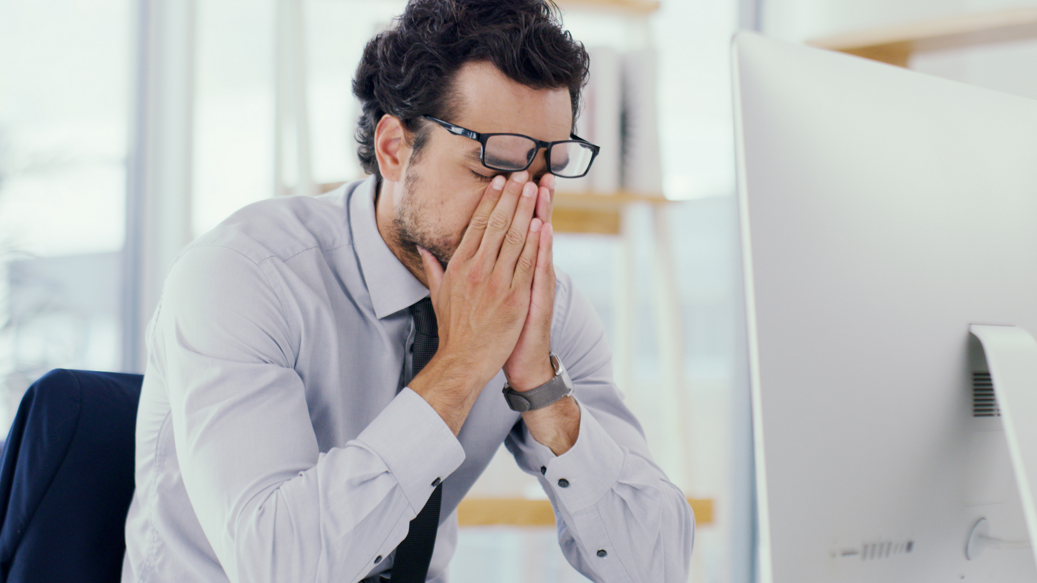 A man with dark hair sitting in front of a computer at a desk with his face in his hands as if tired and frustrated