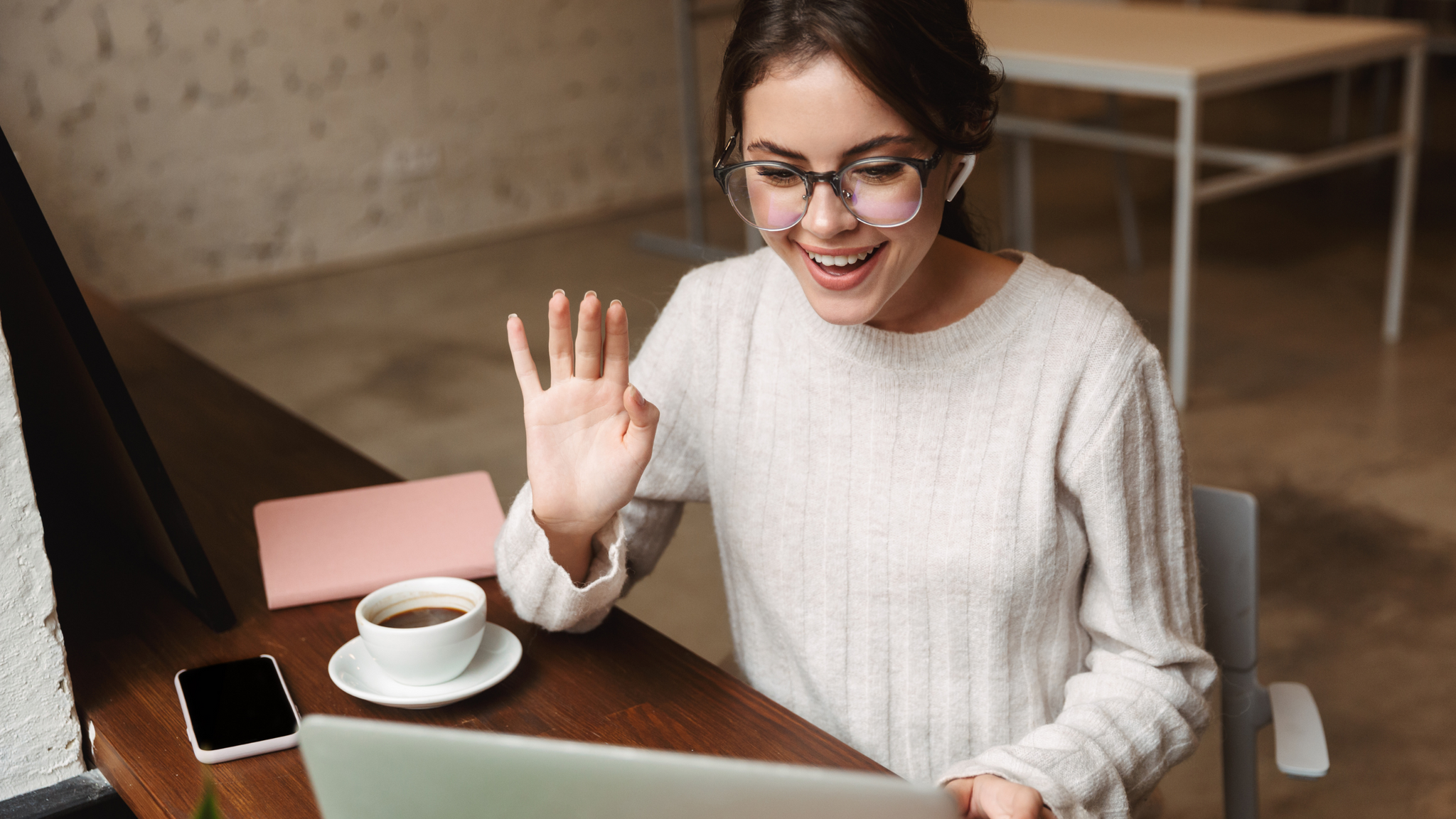 Woman with glasses waving at a computer screen as if saying hi on a video call