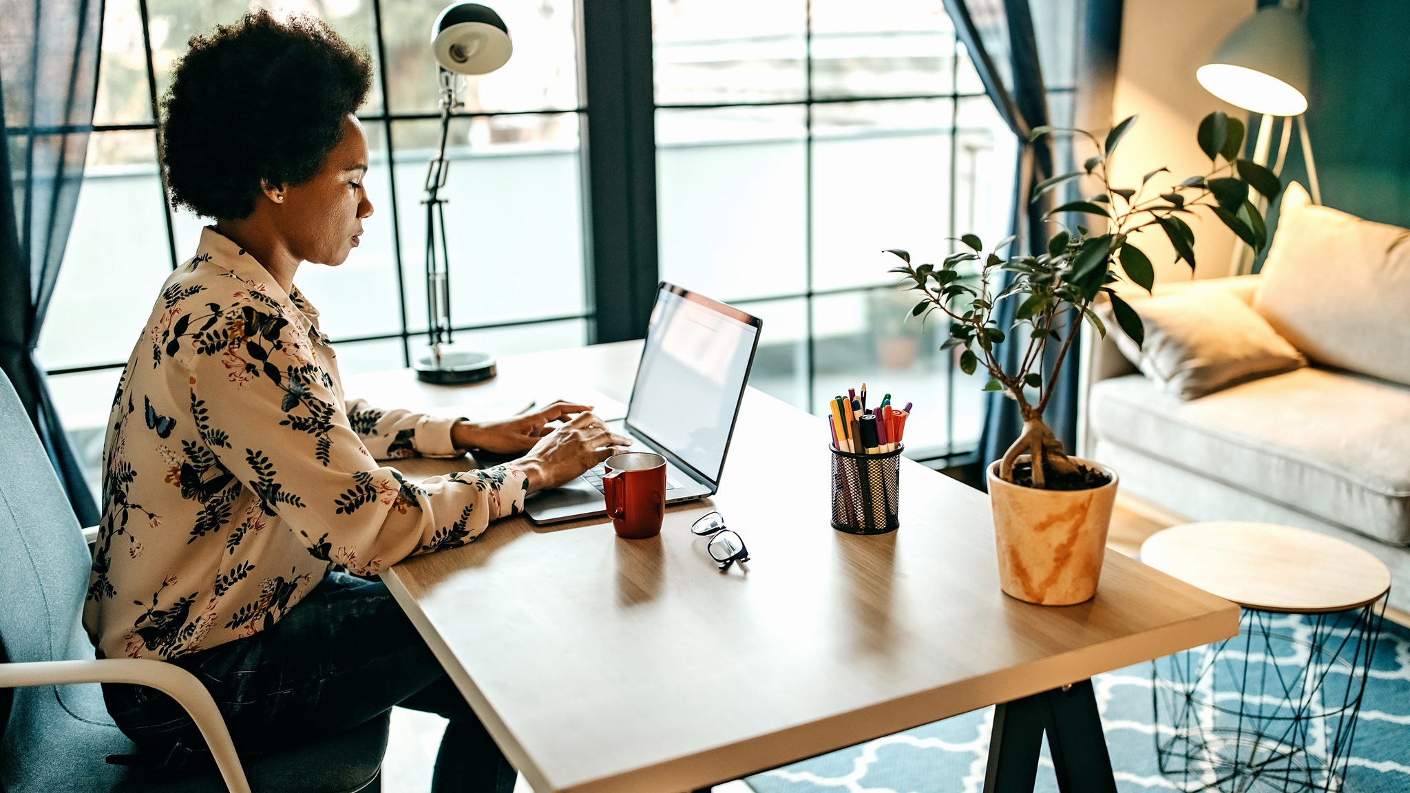 a woman sitting at a small table with a plant working on a laptop