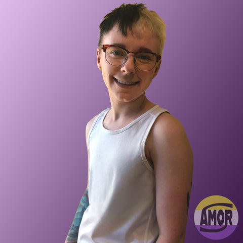 Max, a white non-binary trans person is pictured from the waist up wearing a size S, light grey racerback chest binder, that's not visible, underneath a white sports tank. 