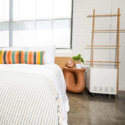 EnviroKlenz Mobile Air System in a bright bedroom next to a white bed with striped bedding and a colorful pillow, with a bamboo ladder and a terracotta side table featuring a salt lamp and potted plant